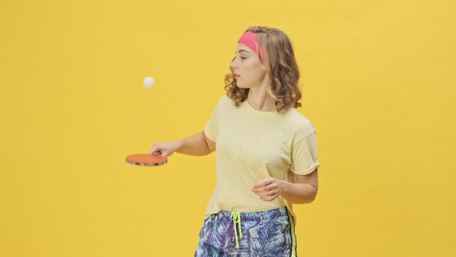 A happy smiling young athletic woman in sportswear is playing ping-pong isolated over a yellow background in studio