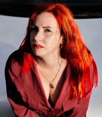 A young girl with red hair and a red shirt with a neckline poses by car.