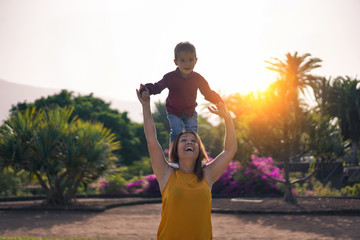 Young mother carries her son on shoulders and laughs - mother and son day in park and have fun -...