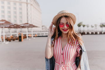Outdoor portrait of pleasant stylish girl in vintage hat standing on city background. Good-humoured female model in pink sunglasses enjoying warm spring day.