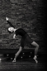 Beautiful young female classical ballet dancer on pointe shoes wearing a black leotard and skirt on a brick background