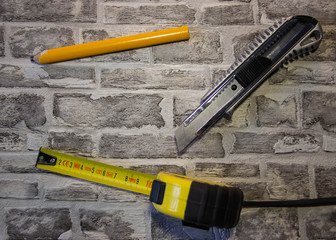 Construction yellow tape measure and yellow pencil, stationery knife on a brick wall background.