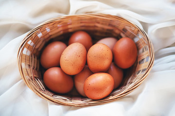 There are brown chicken eggs in a wooden wicker basket. The basket lies in the middle of a soft, thin white cloth.