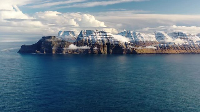 Aerial, pan, drone shot of a snowy mountain ridge,surrounded by sea, during sunrise, in the fjords of Faroe islands, Denmark