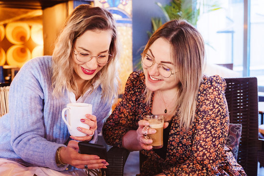 Cheerful women drinking coffee at a terrace on spring holidays and calling a friend with smartphone. Young girlfriends having coffee and using technology at a restaurant. Travel and leisure concept.