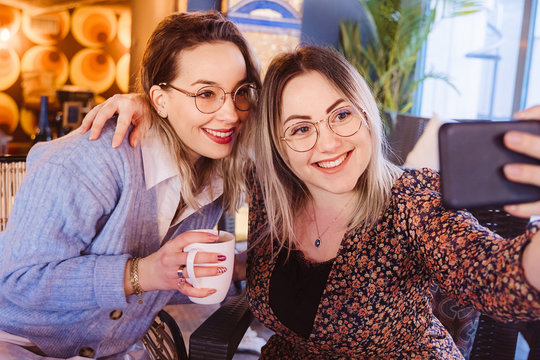 Cheerful women making a video call with a friend with smartphone. Young girlfriends having coffee and using technology at a restaurant. Travel and leisure concept. Technological lifestyle.
