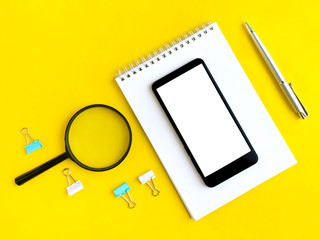 Office flat lay with smartphone, notepad, magnifier, pen and clips on yellow background. Job searching. E-learning. Top view