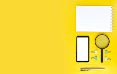 Creative office flatlay with notepad, smart phone, loupe and stationery on yellow background. Job searching or e-learnong concept. Modern minimalist style. Banner with copyspace.