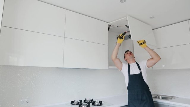 Man dressed in uniform assembling furniture in new house. Carpenter repair and assembling cabinet at the modern kitchen of white color. Slider shot