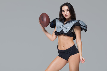 Young sporty brunette wearing sexy uniform of American football player posing with ball looking at camera isolated on grey background