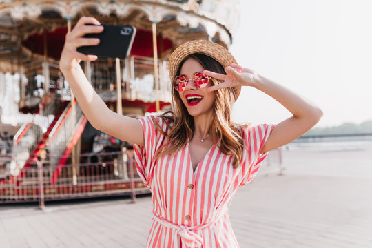 Wonderful slim lady in hat using smartphone for selfie in summer day. Outdoor photo of ecstatic girl in striped dress taking picture of herself in amusement park.