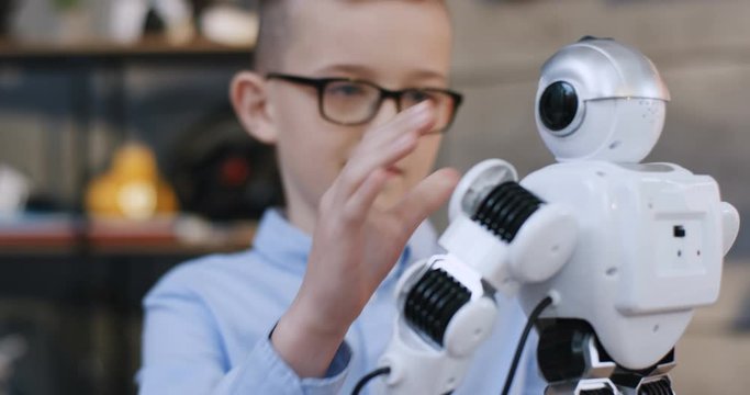 Close up of the cute little Caucasian teen boy in glasses holding a toy robot and playing with it. Indoor.