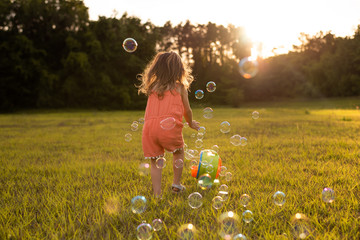 Little girl in orange romper catching soap bubbles on grass in a field at sunset. 