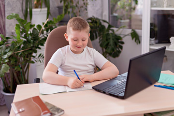 A boy with blond hair in a white T-shirt at the table and does homework on a laptop. Distance learning at home, homework during the epidemic.