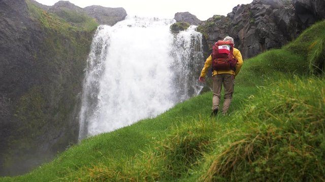 Man hiking up on grassy hill, The Dynjandi waterfall in background, overcast, summer day, in Iceland - slow motion shot