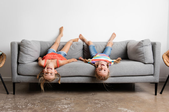 Two smiling little girls lying upside down on sofa at home