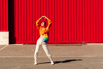 Lesbian woman shaping heart, with colorful bag in front of a red wall