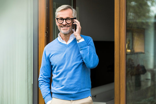 Smiling senior man with grey hair standing in front of his modern design home talking on the phone - Powered by Adobe
