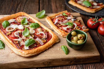 Homemade puff pastry pizza with bacon slices, tomatoes, green olives, cheese and onion, topped with...