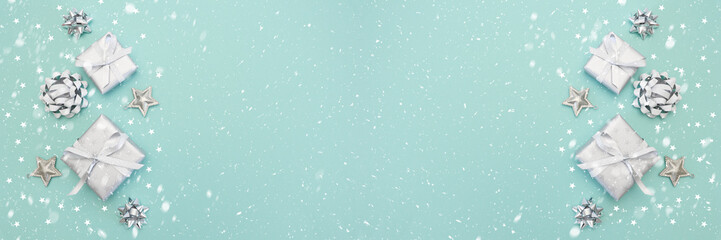 Banner of gifts or present boxes in silver with silver bows and tree ball ornaments and little silver stars, table top view, snowflakes. Composition for Christmas. Turquoise pastel background.