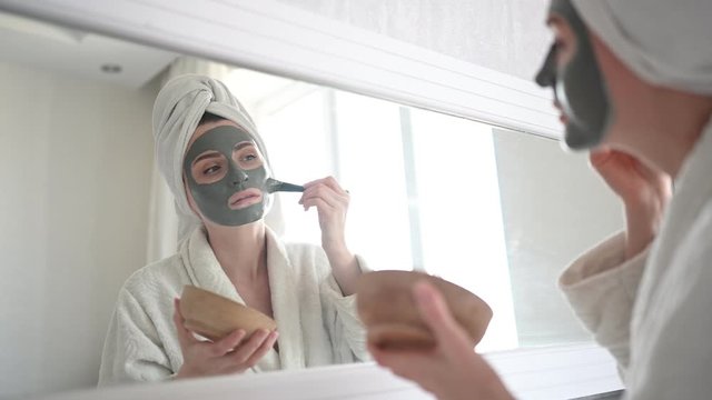 Cosmetology, skin care, face treatment, spa, natural beauty concept. Beautiful smiling woman at home in bathrobe with a towel applying face clay mask against acne to rejuvenate problem skin