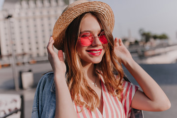 Lovely blithesome girl in pink glasses posing with gently smile. Outdoor shot of stylish white woman touching her straw hat on city background.