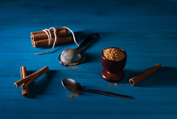 Ground cinnamon powder in wooden cup, a pack of cinnamon sticks, a little spoon and metal sifter on...
