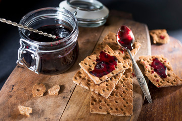 Open jar with blackcurrant jam on a wooden cutting board. Dry biscuits spread with jam, crumbs and...