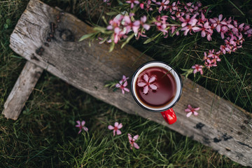 Fototapeta na wymiar Tea party on the street. Tea in a red mug stands on a barn Board on the green grass. Next to the branches with pink flowers. A flower floats in the Cup.