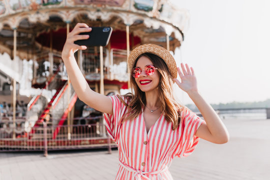 Lovable young woman in summer dress using phone for selfie. Outdoor shot of laughing girl in straw hat holding smartphone and taking picture of herself near amusement park.