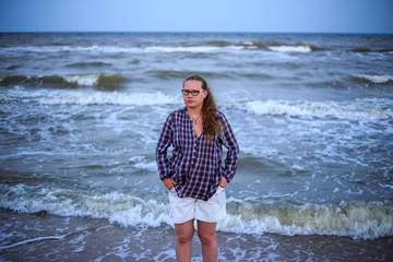 A beautiful woman with long hair stands against the backdrop of a raging sea. A woman is wearing white shorts and a blue shirt. Stormy sea in the background.