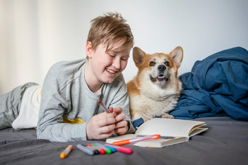 Portrait of happy boy lying on bed with his dog using felt tip pens and sketch book