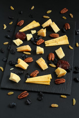 Pieces of parmesan cheese and gouda cheese with fenugreek, blue and gold raisins and dried apricots on black stone plate on black table. Closeup