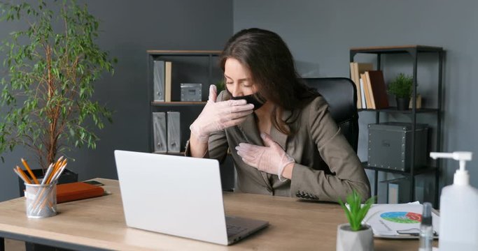 Young ill Caucasian businesswoman in mask and rubber gloves typing on keyboard and working on laptop computer. Unhealthy tired female office worker caughing and stretching neck. Coronavirus.