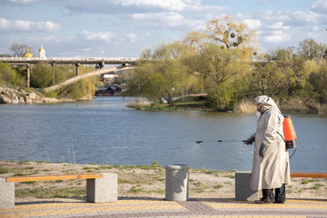 Fototapeta premium Bila Tserkva, Ukraine - April 20, 2020: A man in a gray coat treats the area with a cleaning solution. Shops, trash cans processing from Covid-19. The Covid-19 Epidemic. Pandemic. Beach. River.