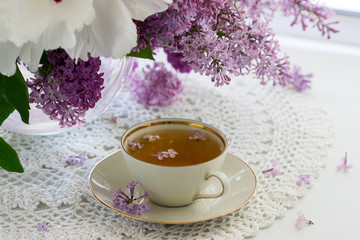A cup of chamomile tea in a porcelain cup and a bouquet of lilac with peony on a windowsill.