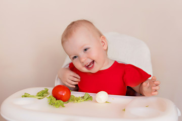 A cheerful baby 10-12 months old eats vegetables. Portrait of a happy girl in a highchair.