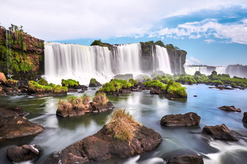 Iguazu waterfalls in Argentina, view from Devil's Mouth. Panoramic view of many majestic powerful water cascades with mist and clouds. Panoramic image of Iguazu valley with grass and stones in water.