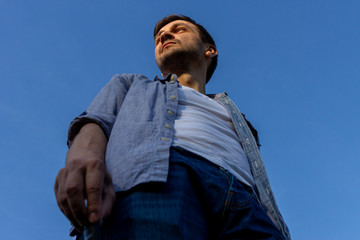 a young man wearing jeans, a white T-shirt and a blue shirt stands in a field against a blue cloudless sky, bottom view. © dimakig