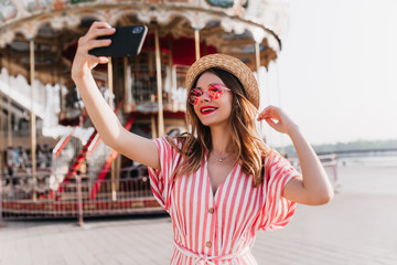 Blithesome female model in striped attire posing near carousel in straw hat. Outdoor shot of fashionable caucasian girl using smartphone for selfie in amusement park. - obrazy, fototapety, plakaty