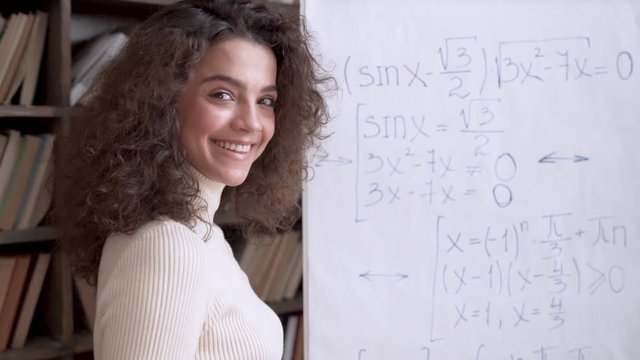 Smiling hispanic young woman math class teacher, tutor standing at whiteboard teaching mathematical equation looking at camera. Happy female university college student learning in classroom. Portrait.