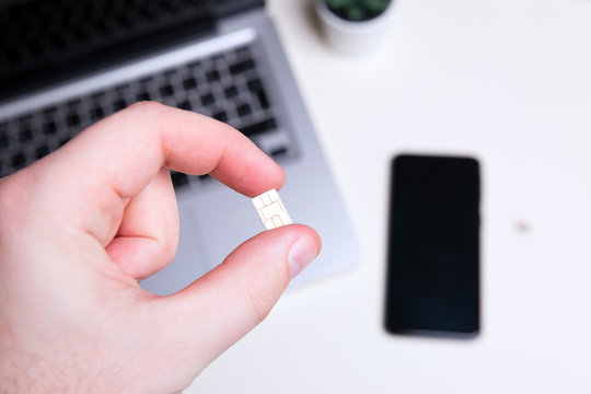 Sim card in a man&rsquo;s hand, phone and computer in the background
