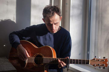 Obraz premium Guy with a guitar in the kitchen. Natural light comes to the musician from the window. Lesson playing the guitar. Unknown talented musician. Playing an acoustic guitar. Brutal man shows skill.