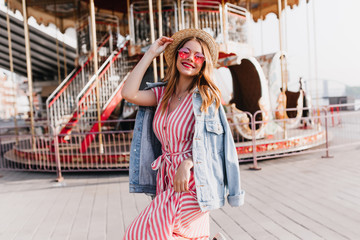 Wonderful girl in straw hat and denim jacket posing near carousel. Outdoor photo of laughing beautiful lady chilling in amusement park.