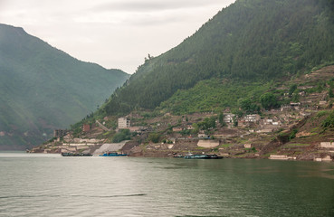 Fototapeta premium Shengli Street, China - May 6, 2010: Xiling gorge on Yangtze River. Rows of houses near green water shoreline on brown dirt slope beneath green tree covered mountains. Some vessels anchored.