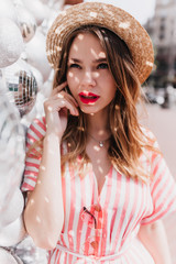 Fascinating girl in retro straw hat posing on city background. Outdoor photo of elegant caucasian woman in striped dress standing near disco balls.