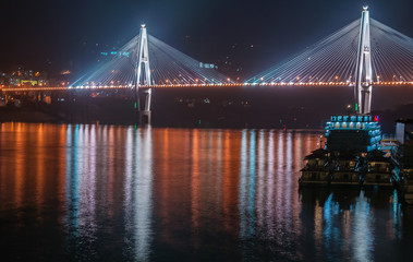 Xinling, China - May 6, 2010: Xiling gorge on Yangtze River. Night picture of Badong Bridge with its 2 pylons and fully lighted in 3 colors. Boats on side. City lights.