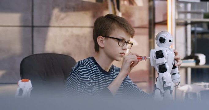 View from far and blurred point on the cute small Caucasian schoolboy in glasses twisting nuts in the white toy robot with a screwdriver while repairing it at the technological workshop.