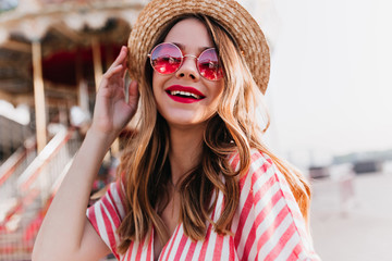 Dreamy girl in round pink glasses posing with smile in amusement park. Outdoor portrait of romantic blonde lady wears straw hat during summer photoshoot.