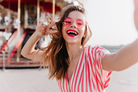 Adorable girl in stylish sunglasses posing with inspired face expression beside carousel. Outdoor photo of blissful white woman making selfie in amusement park.